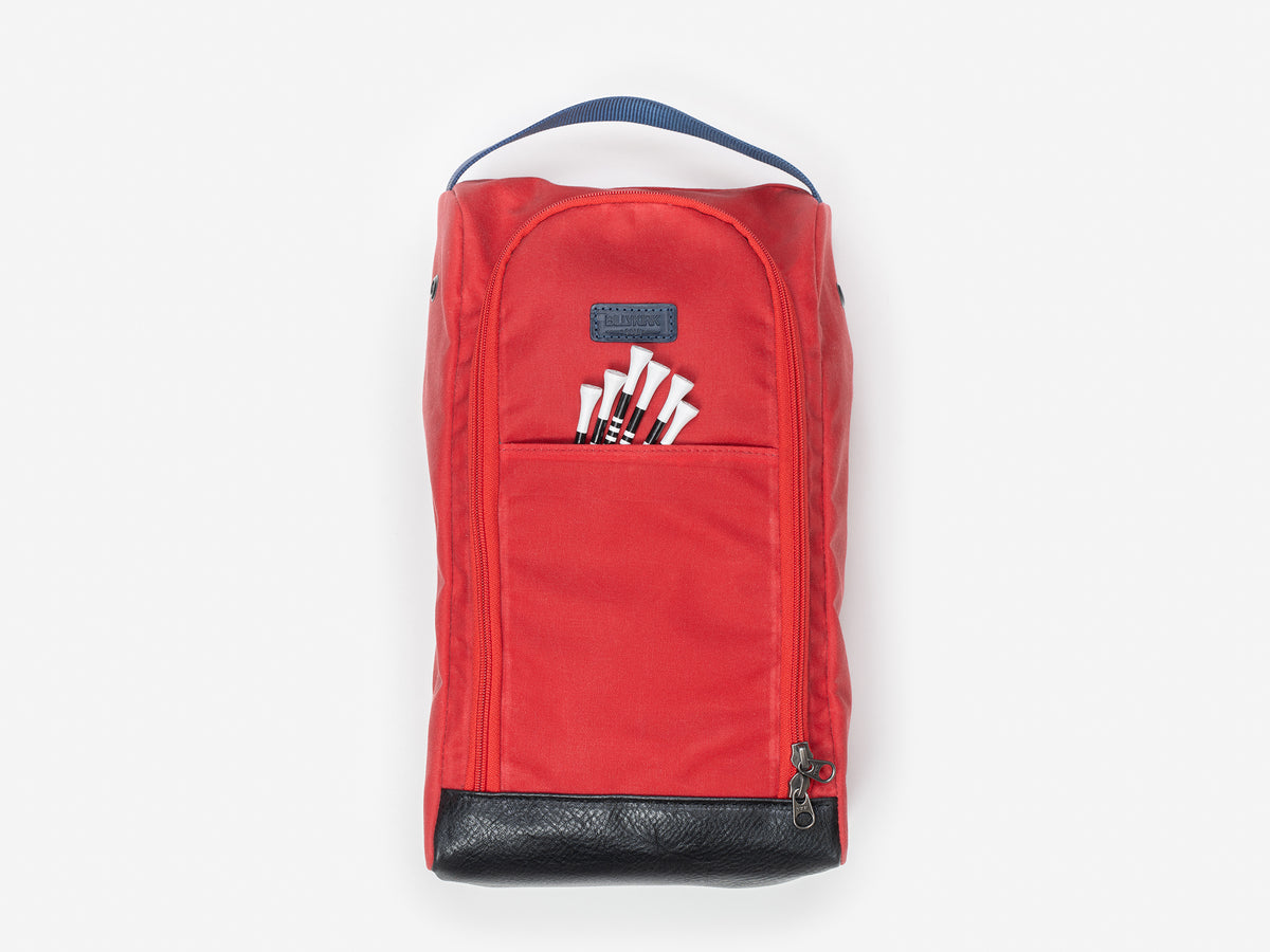 A red fabric shoe bag with a black handle on top, a front zipper pocket, a front flap with six white tees, and a black base, displayed against a plain light background.