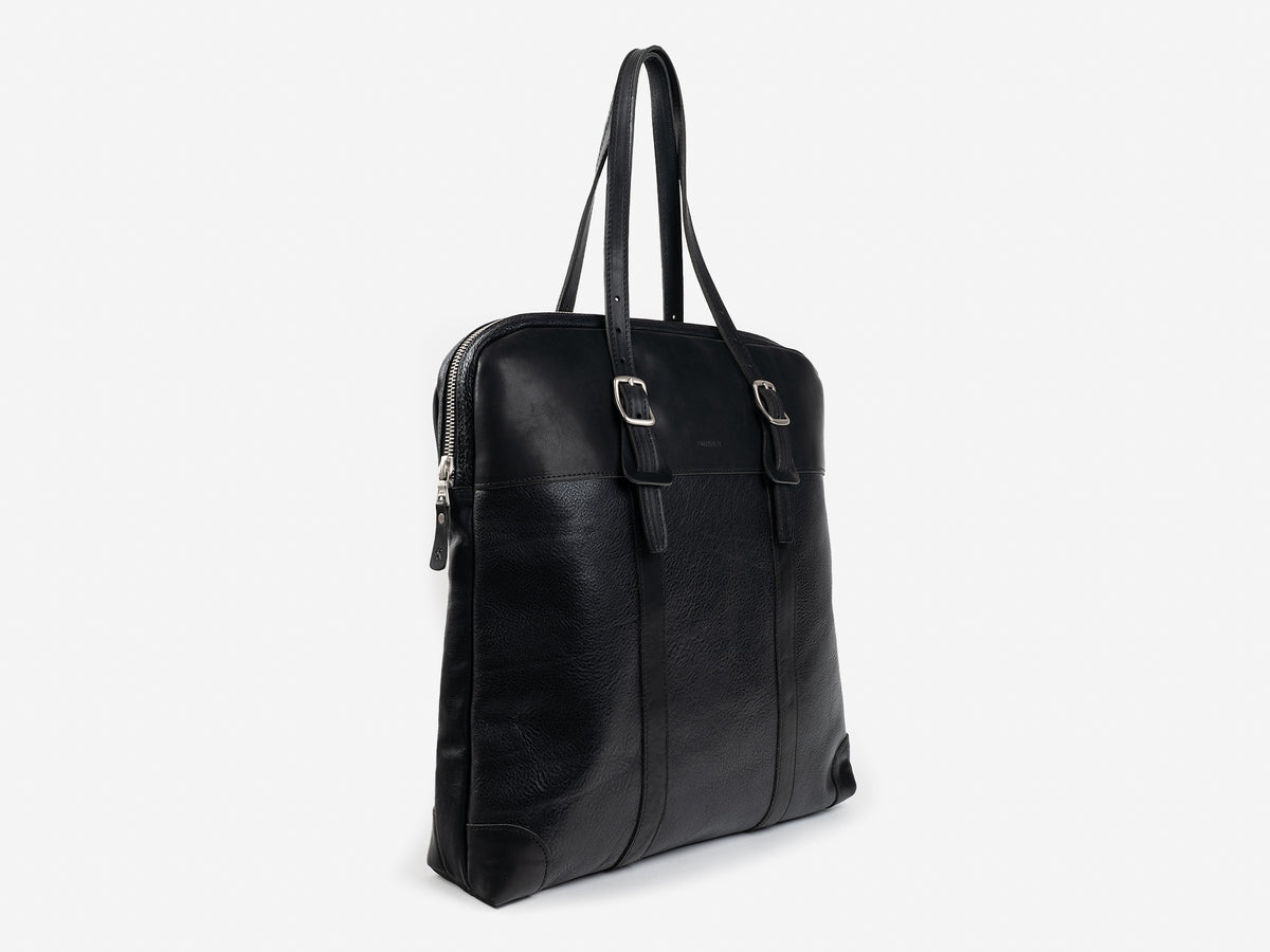 A black leather tote bag with two shoulder straps, a zipper closure, and metal buckle accents, shown standing upright on a plain white background.
