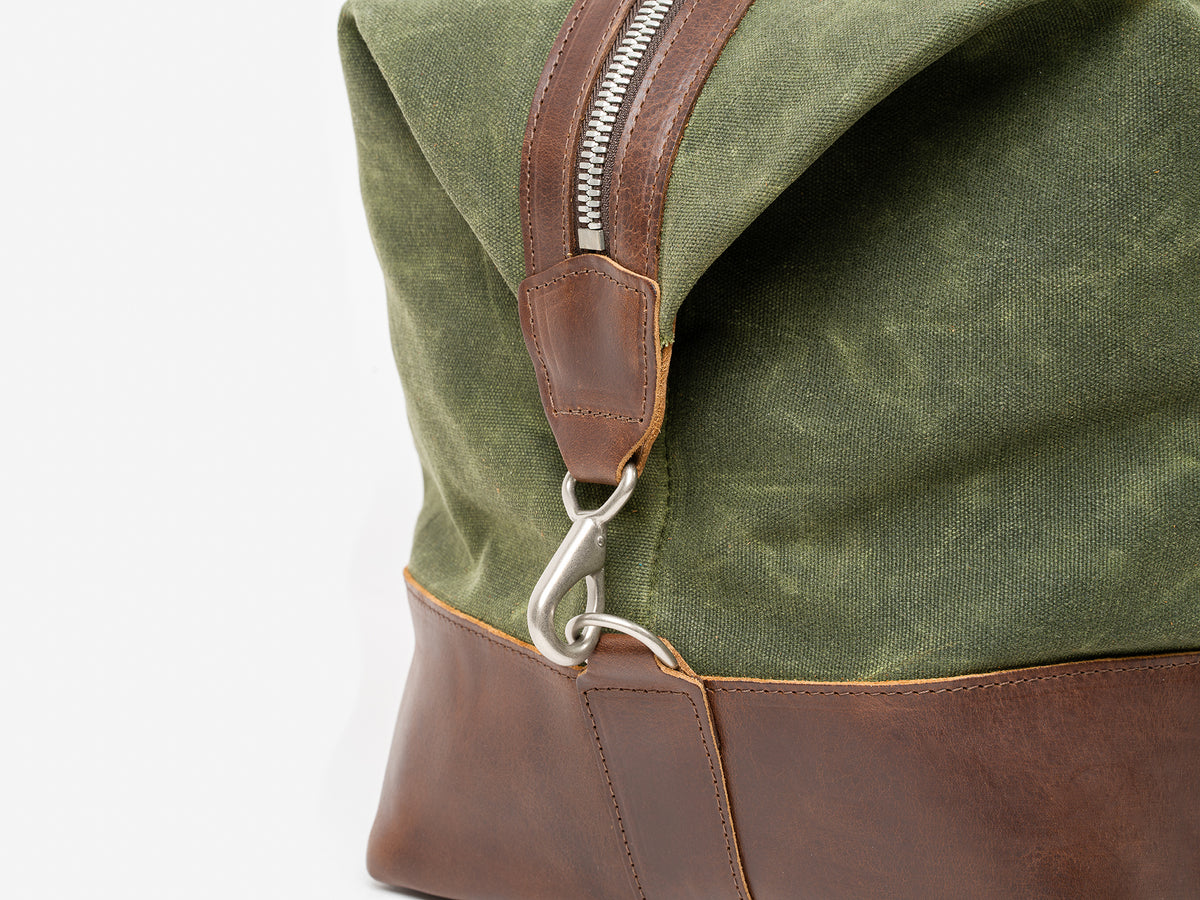 Close-up of a Green and Brown  leather bag, featuring a silver zipper and metal clasp attached to a leather strap. The image highlights the textures and details of the materials.