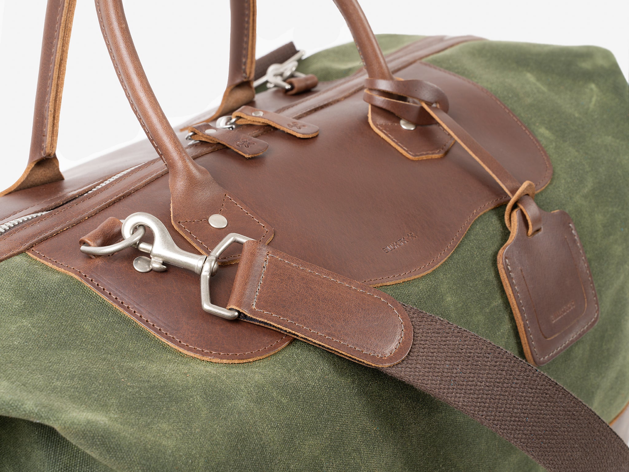 A close-up of a green canvas duffel bag with brown leather straps, trims, and a luggage tag. The bag features silver metal hardware and a sturdy zipper, highlighting its high-quality craftsmanship.