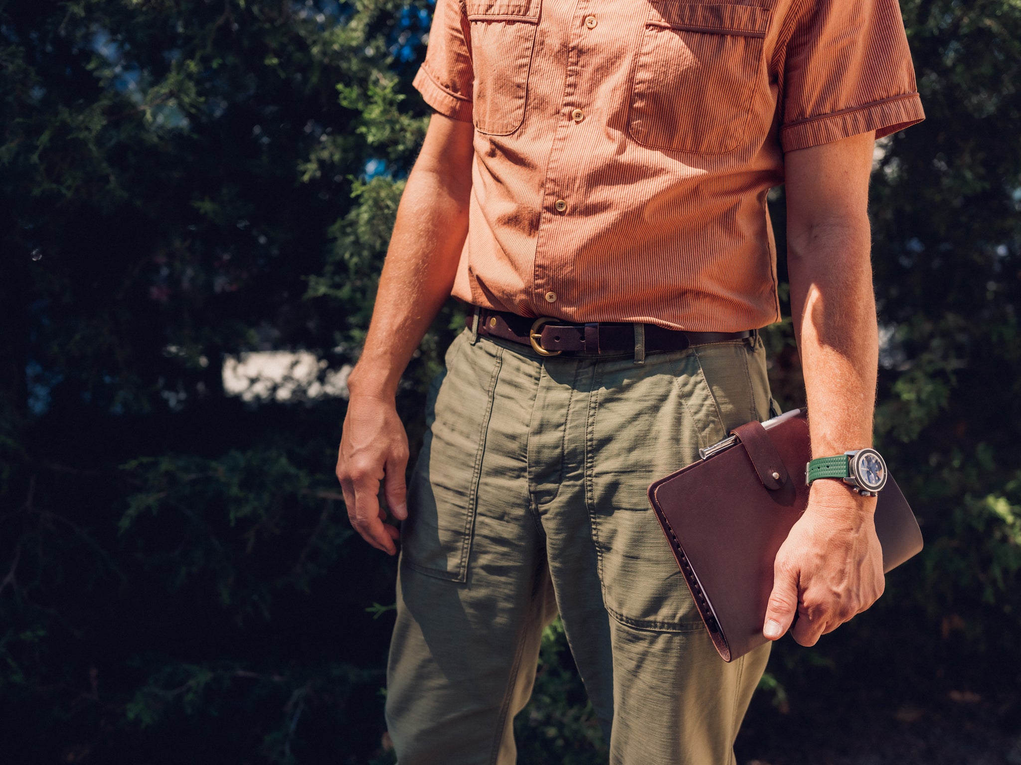 A person wearing an orange short-sleeve shirt and green pants stands outdoors, holding a brown leather folder in one hand. The background has green foliage and the person is partially visible from shoulders down.