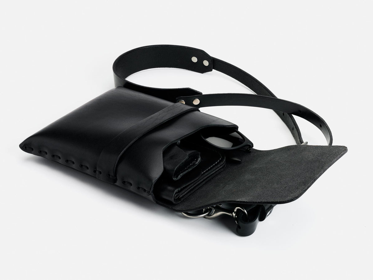 A black leather crossbody bag with an open flap, showing a wallet, sunglasses, and keys inside. The bag has a long adjustable strap and metal studs along the edges.