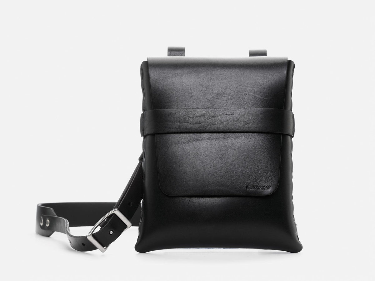 A minimalist black leather crossbody bag with a front flap, simple strap, and silver buckle, shown against a plain white background.