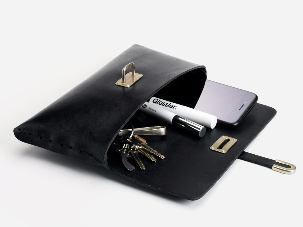 A black leather clutch lies open on a white surface, revealing a smartphone, several keys, and a Glossier-branded tube inside. The clasp and hardware are gold-colored.