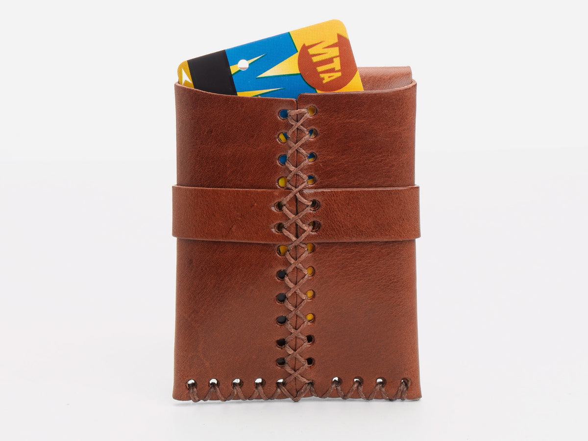 A tan leather wallet with visible stitching holds a New York City MetroCard,  partially sticking out of the rear wallet on a white background.
