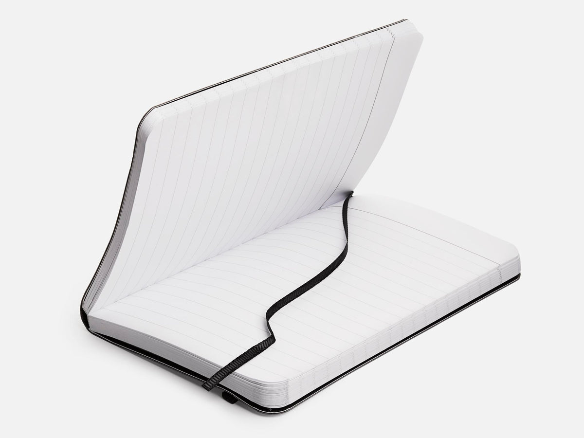 An open, lined notebook with a black cover and a black elastic strap lying across its pages, set against a plain white background.