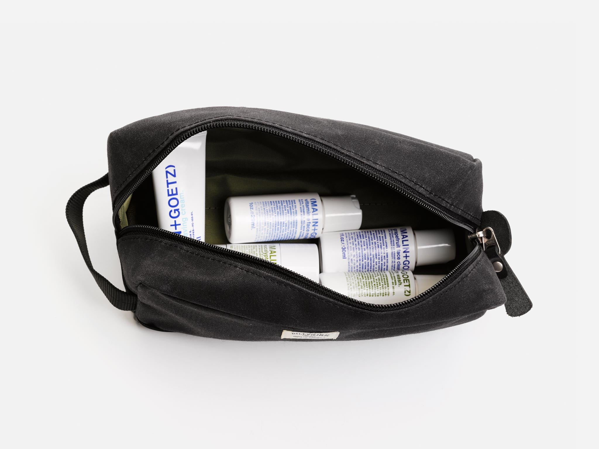 A black waxed canvas toiletry bag unzipped to reveal several Malin+Goetz branded bottles and containers inside, arranged neatly against a white background.