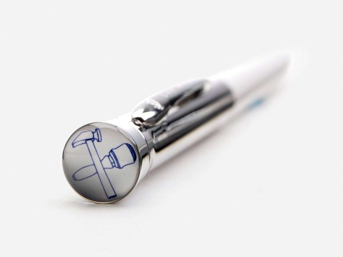 A close-up of a metallic pen resting on a white surface, with a round emblem featuring a blue hammer and pickaxe crossed on the end of the pen.