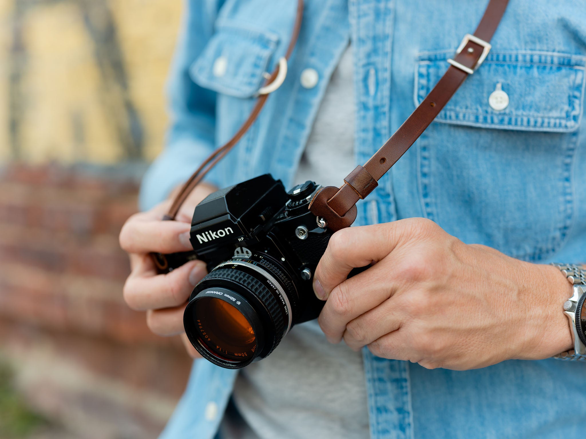 Close-up of a person wearing a denim shirt, holding a Nikon film camera with both hands. The camera hangs from a brown leather strap around their neck. The background is out of focus.