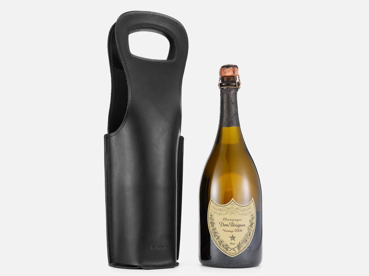 A bottle of Dom Pérignon champagne stands next to a black leather wine bottle holder with a handle, both set against a plain white background.