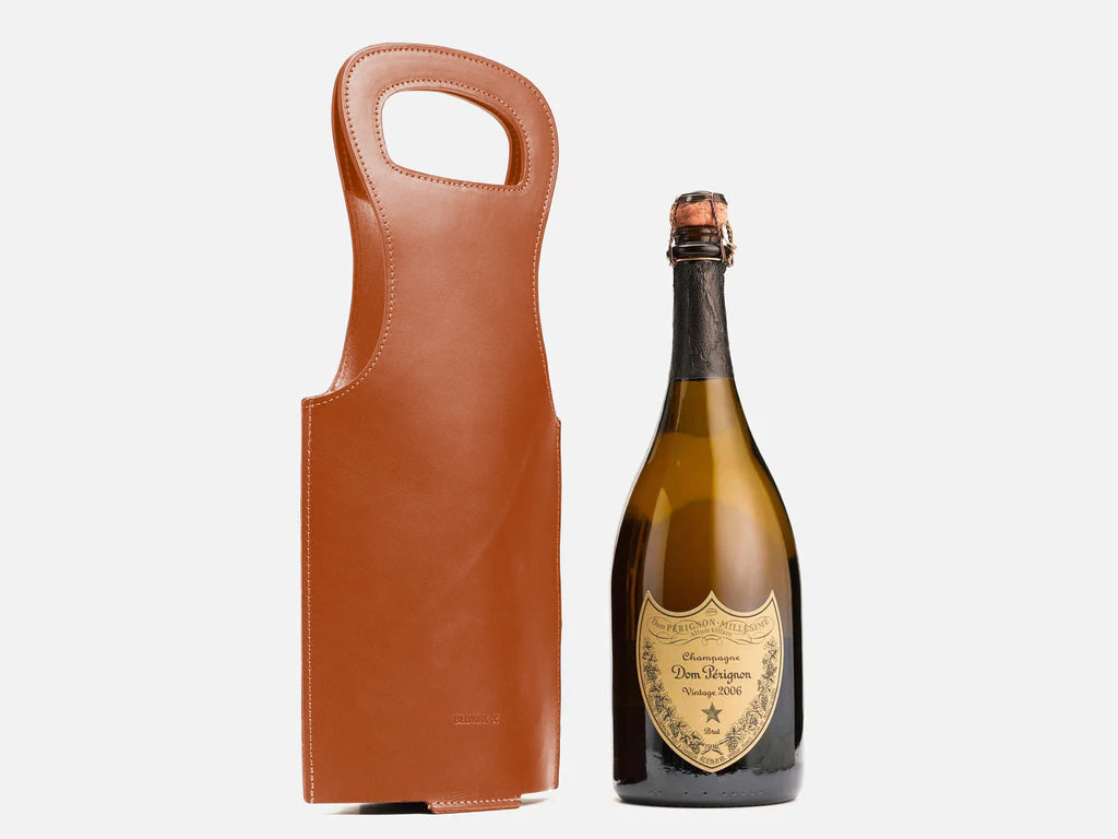 A bottle of Dom Pérignon champagne stands next to a brown leather wine carrier with a handle, both set against a plain white background.