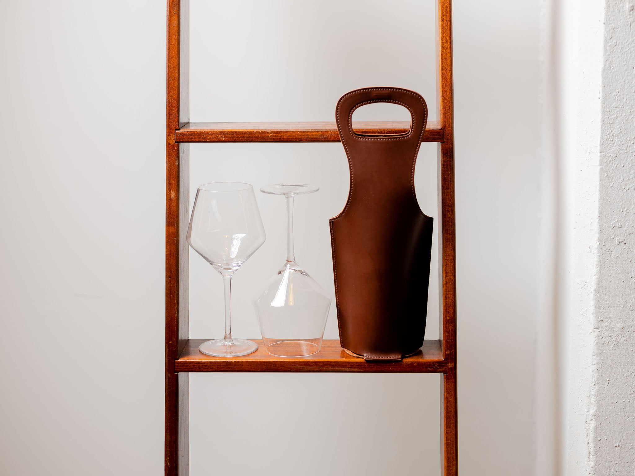 Two wine glasses and a brown leather wine bottle holder are displayed on a wooden shelf against a white wall. One glass is upright and the other is upside down.
