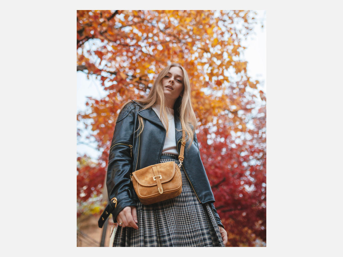 A woman wearing a black leather jacket, plaid skirt, and holding a mustard yellow handbag stands outdoors with vibrant red and orange autumn leaves in the background.
