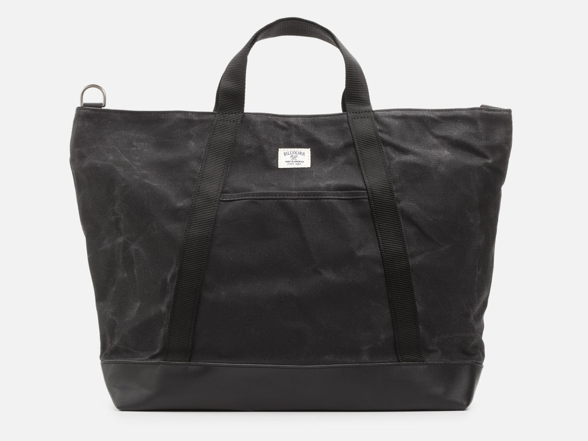 A back view of an black waxed canvas tote bag with 1" nylon black handles with pocket between the handles, and a black leather bottom, set against a plain white background.
