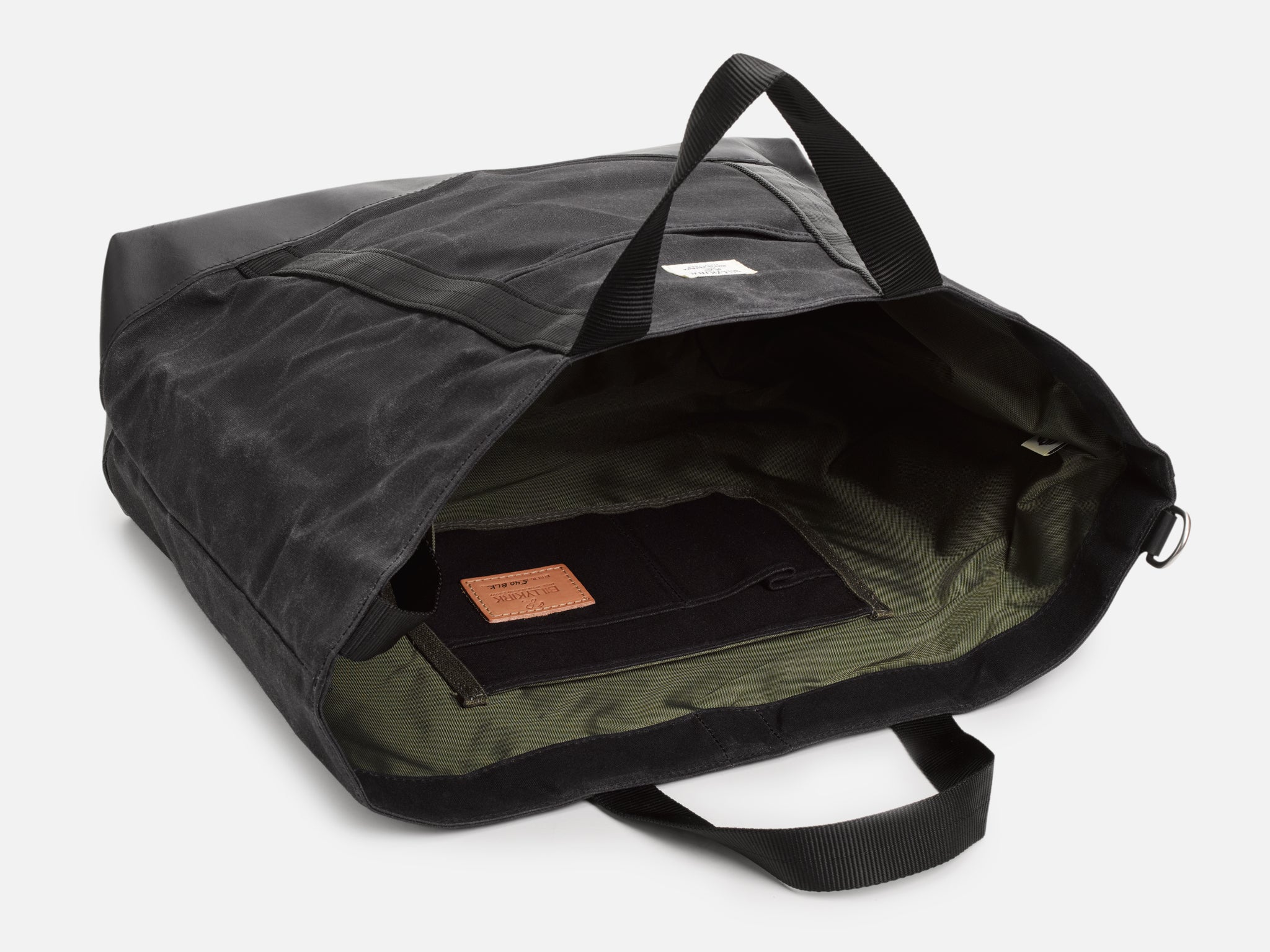 A black tote bag, resting on its back, partially opened, showing olive nylon lining and interior organization pocket with a veg tan leather logo patch.
