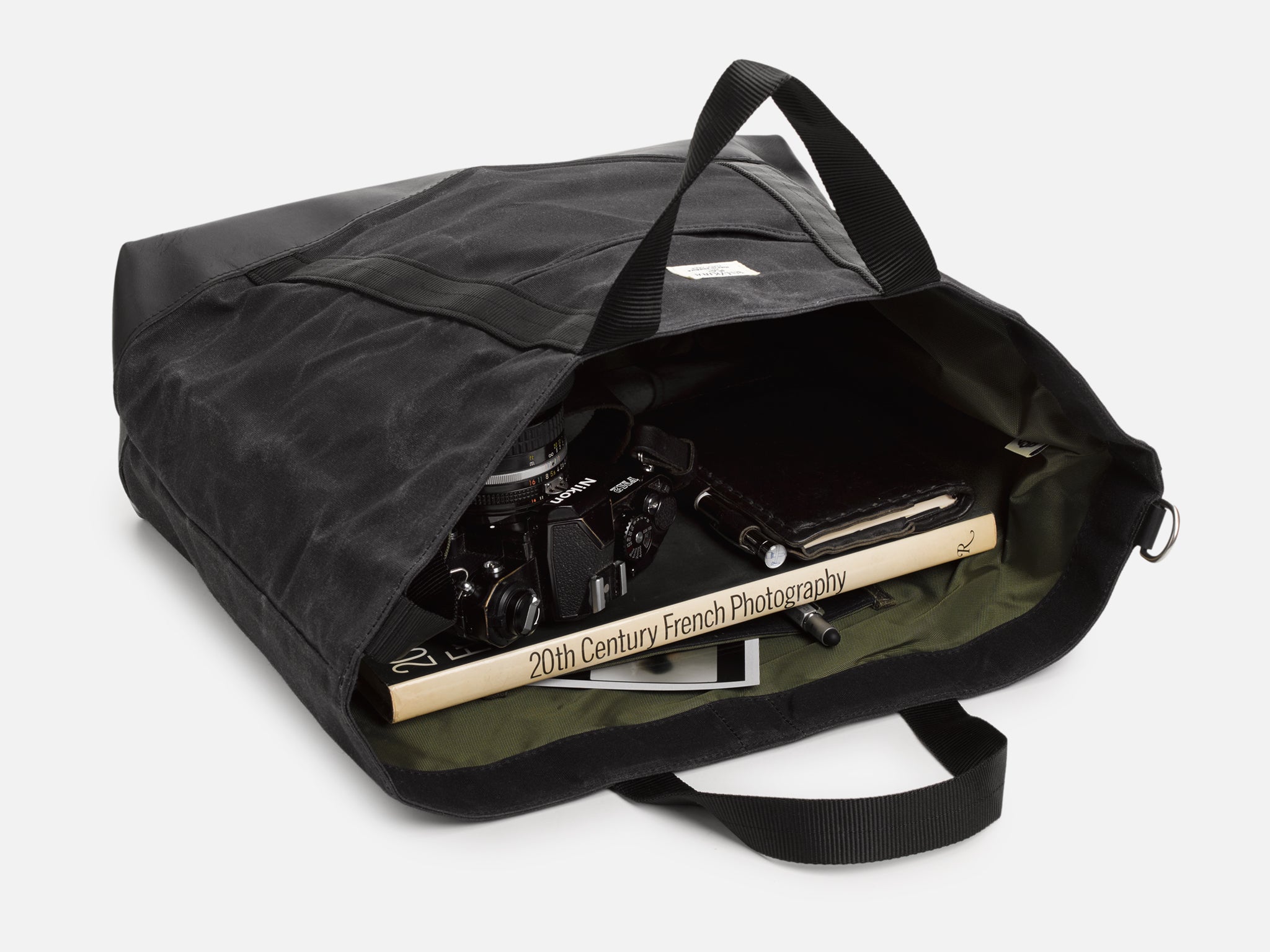 A black tote bag, resting on its back, partially opened, reveals a book titled Hammershoi and Europe, a brown wallet, a tablet, and white earphones neatly inside.