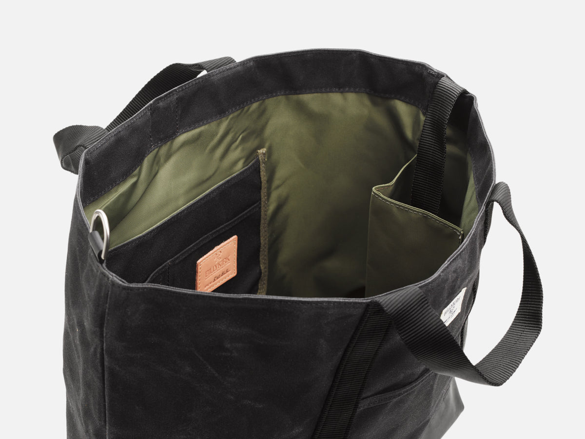 Open black tote bag with black handles, showing an olive green nylon interior with multiple pockets and compartments, including one with a small veg tan leather logo patch and another sized for a bottle or for when the shoulder strap is not in use.