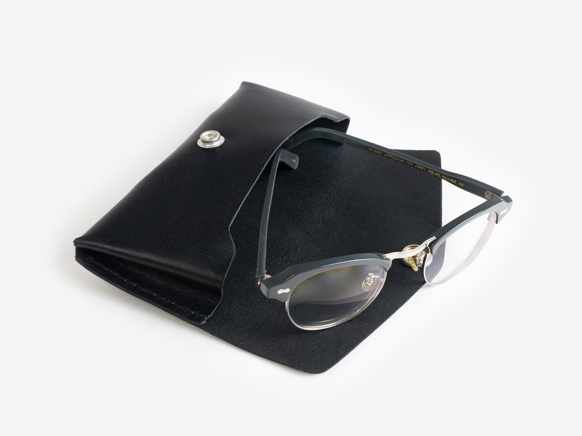 A pair of black-framed eyeglasses with gold accents rests on an open black leather case with a snap closure, set against a white background.