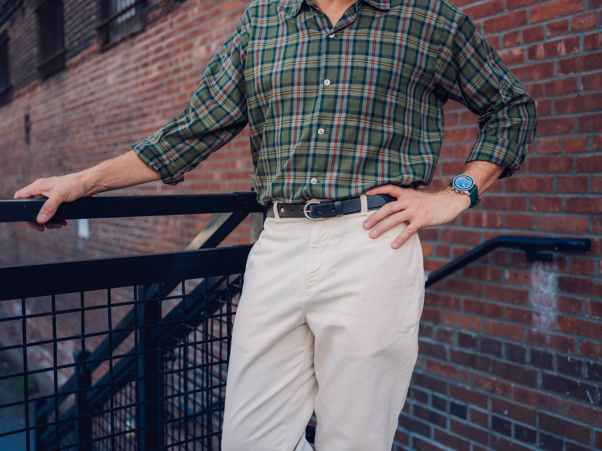 A person wearing a green plaid shirt, beige pants, and a watch stands outside against a brick wall, resting one hand on a black metal railing and the other on their hip. Their face is not visible.