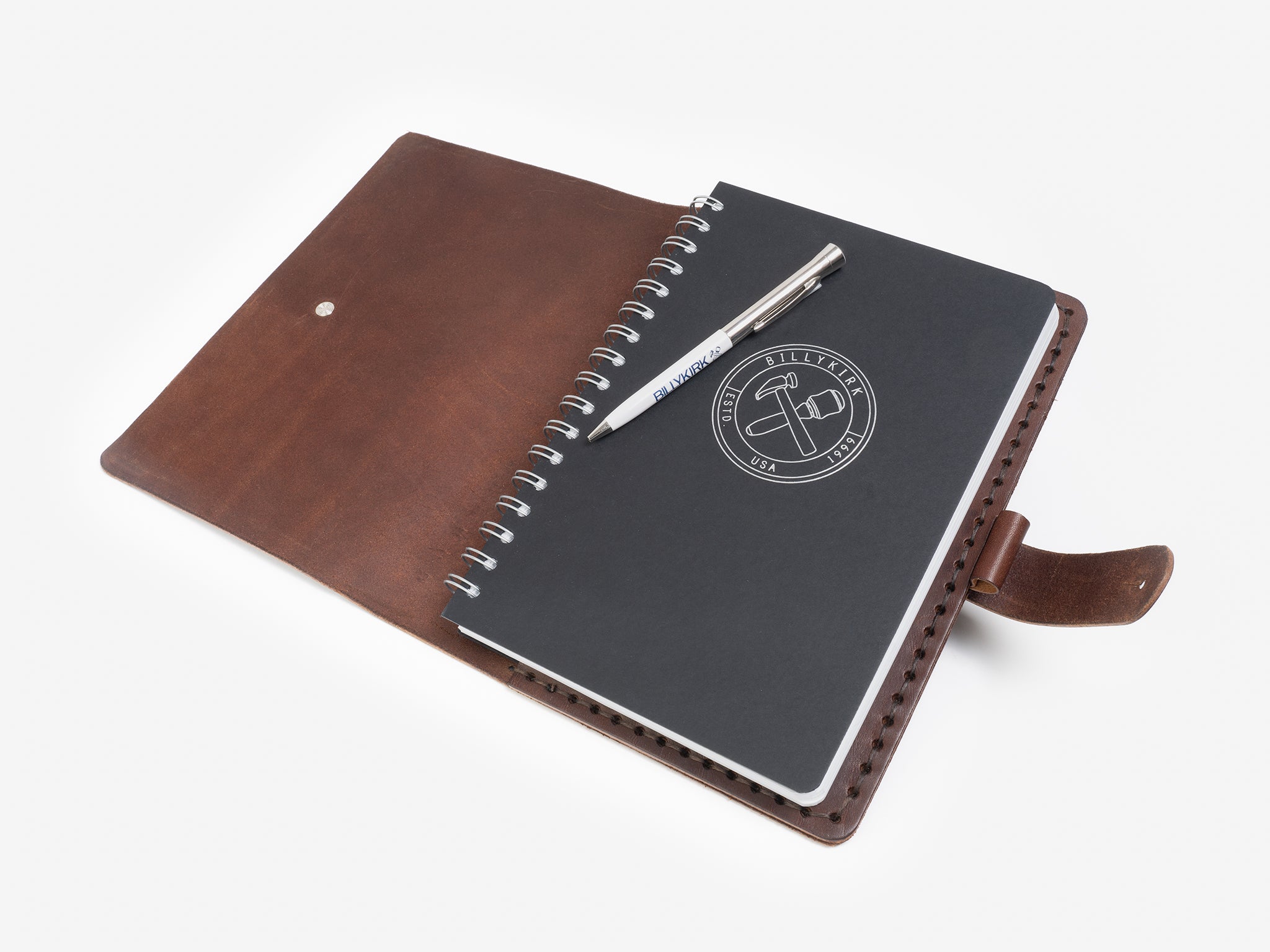 An open brown leather folio with a snap button holds a spiral-bound black notebook showing a logo and a silver pen resting on top, all set against a white background.