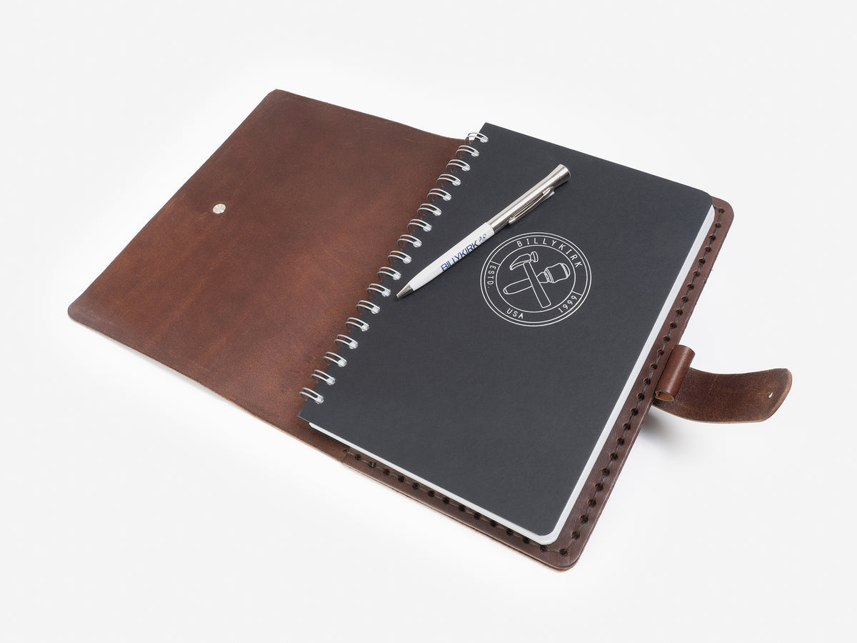 A brown leather folio is open, revealing a black spiral-bound notebook with a logo and a silver pen resting on top, all set against a white background.