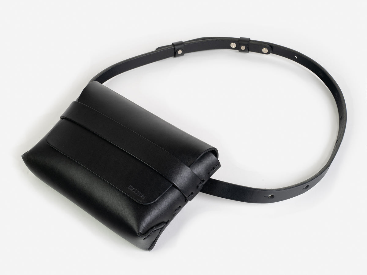 A black leather belt crossbody bag with a smooth finish, featuring a flap closure, subtle embossed branding, and an adjustable strap with silver-tone collar buttons, displayed against a white background.