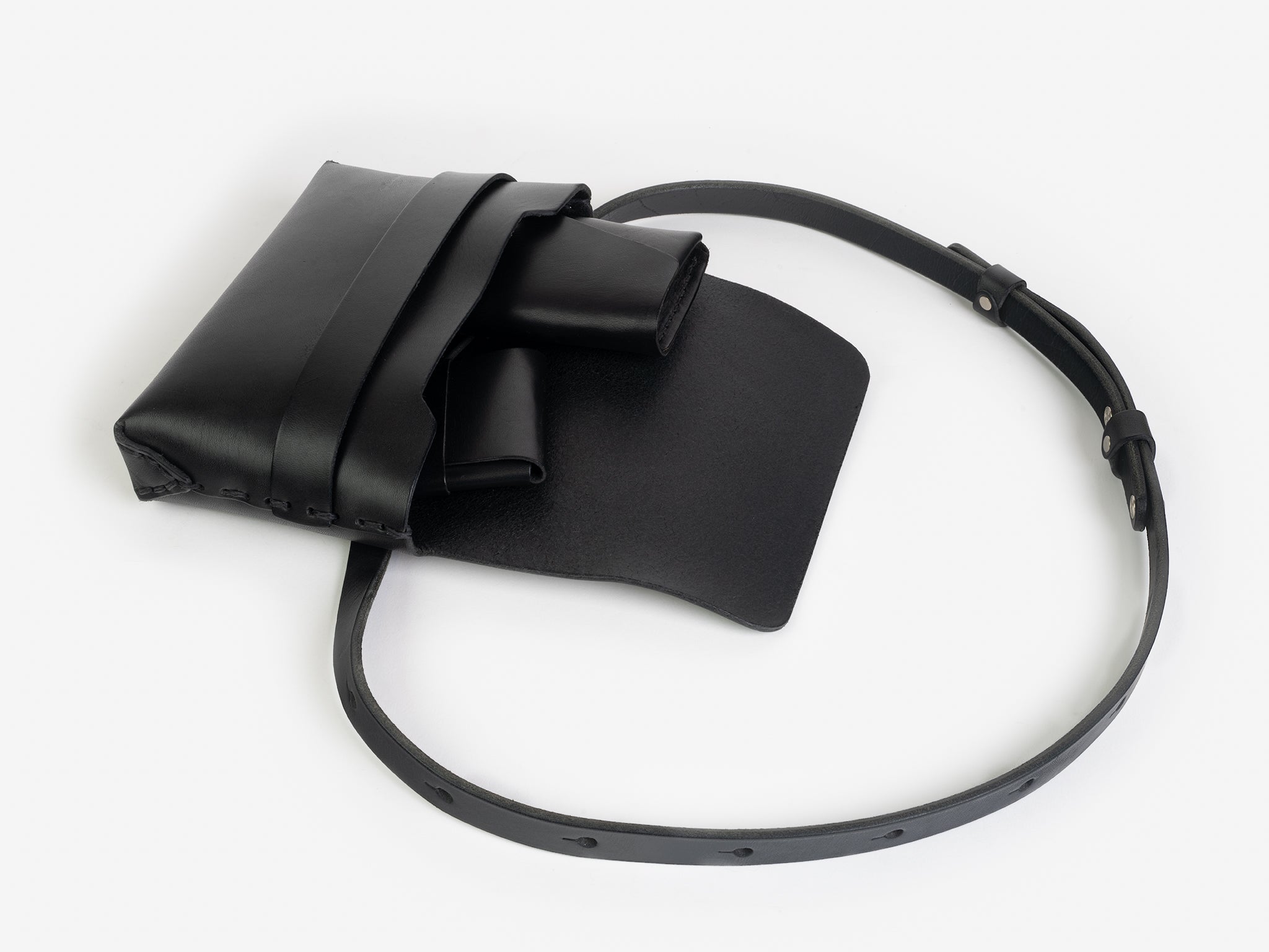 A black leather crossbody bag with a flap open, revealing a black leather card case, and a  black leather sunglasses case inside. The bag has a long adjustable strap and minimalist design, displayed on a white background.