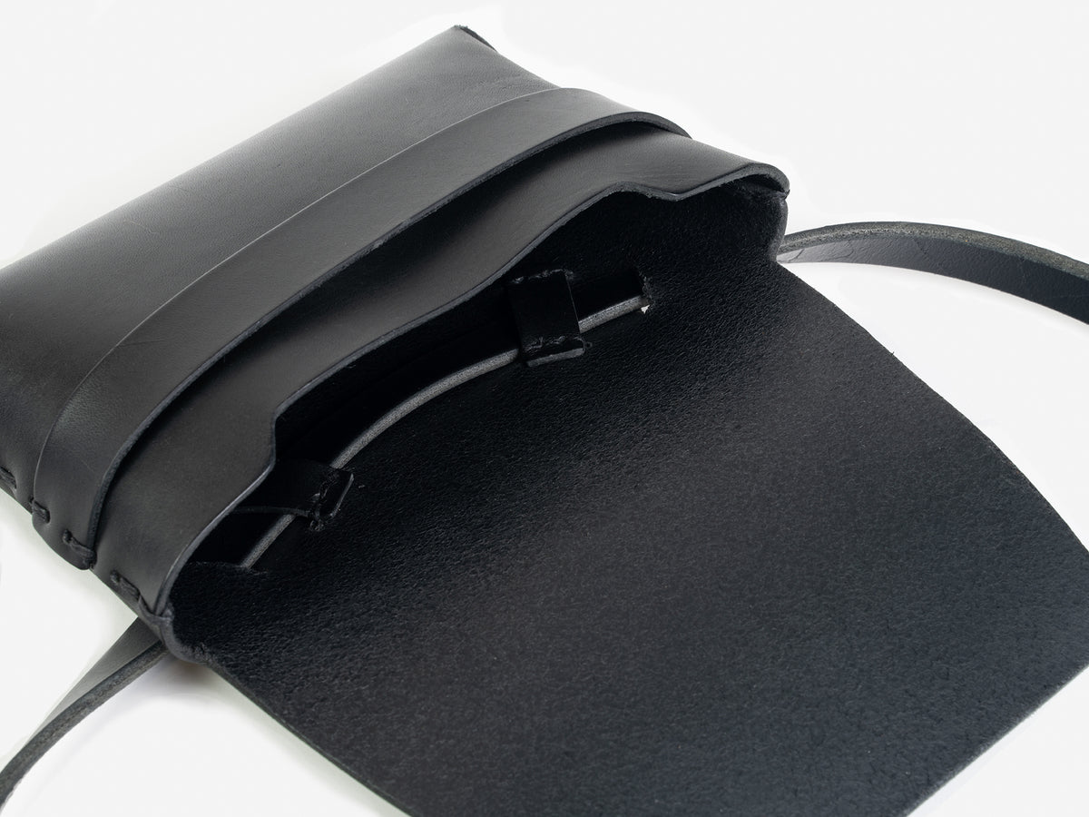 A black leather crossbody bag with a flap open, revealing some hand-stitched details. The bag has a strap and minimalist design, displayed on a white background.