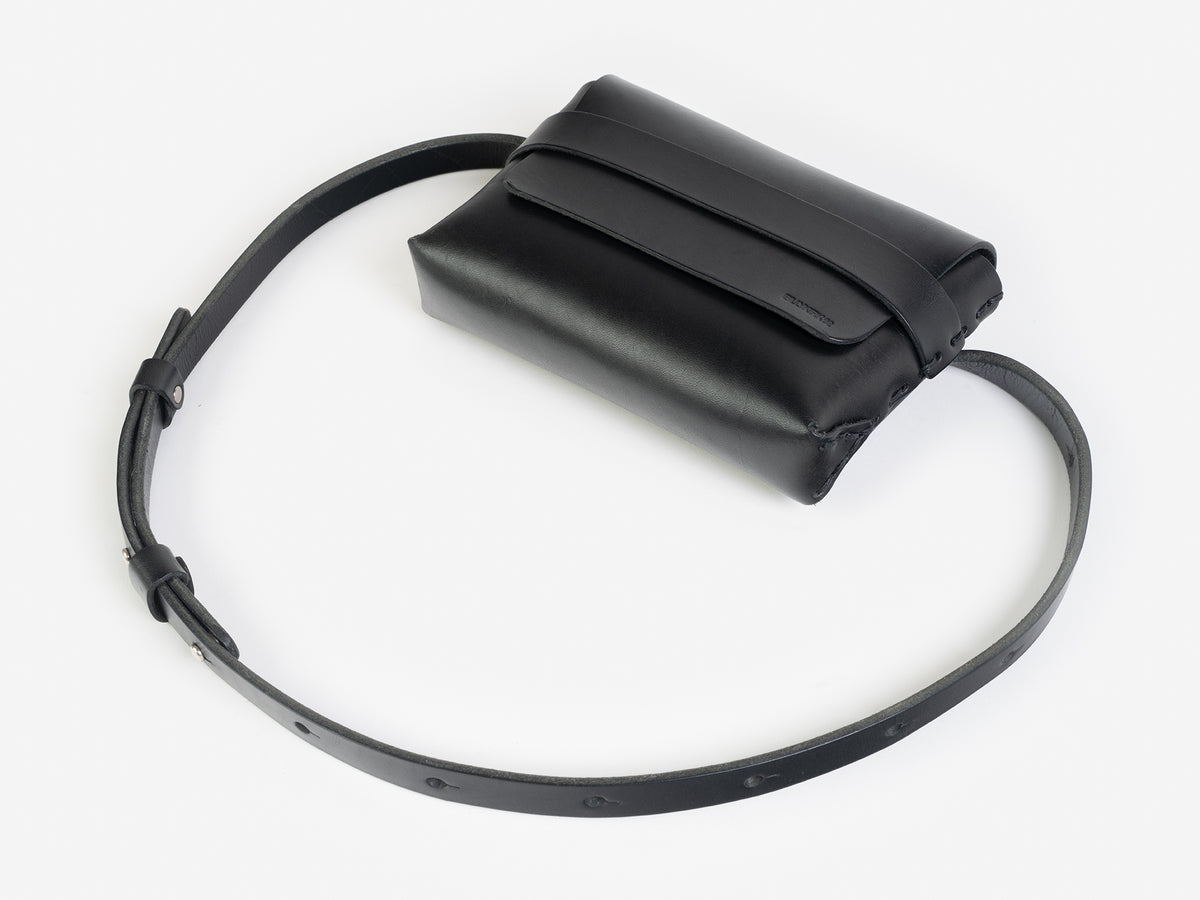 A black leather belt crossbody bag with a smooth finish, featuring a flap closure, subtle embossed branding, and an adjustable strap with silver-tone collar buttons, displayed against a white background.