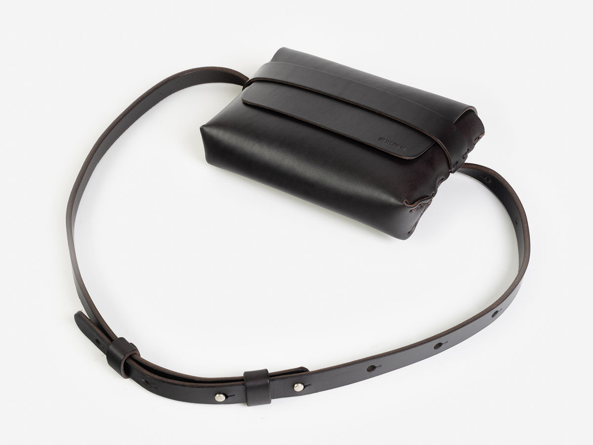 A dark brown leather belt crossbody bag with a smooth finish, featuring a flap closure, subtle embossed branding, and an adjustable strap with silver-tone collar buttons, displayed against a white background.