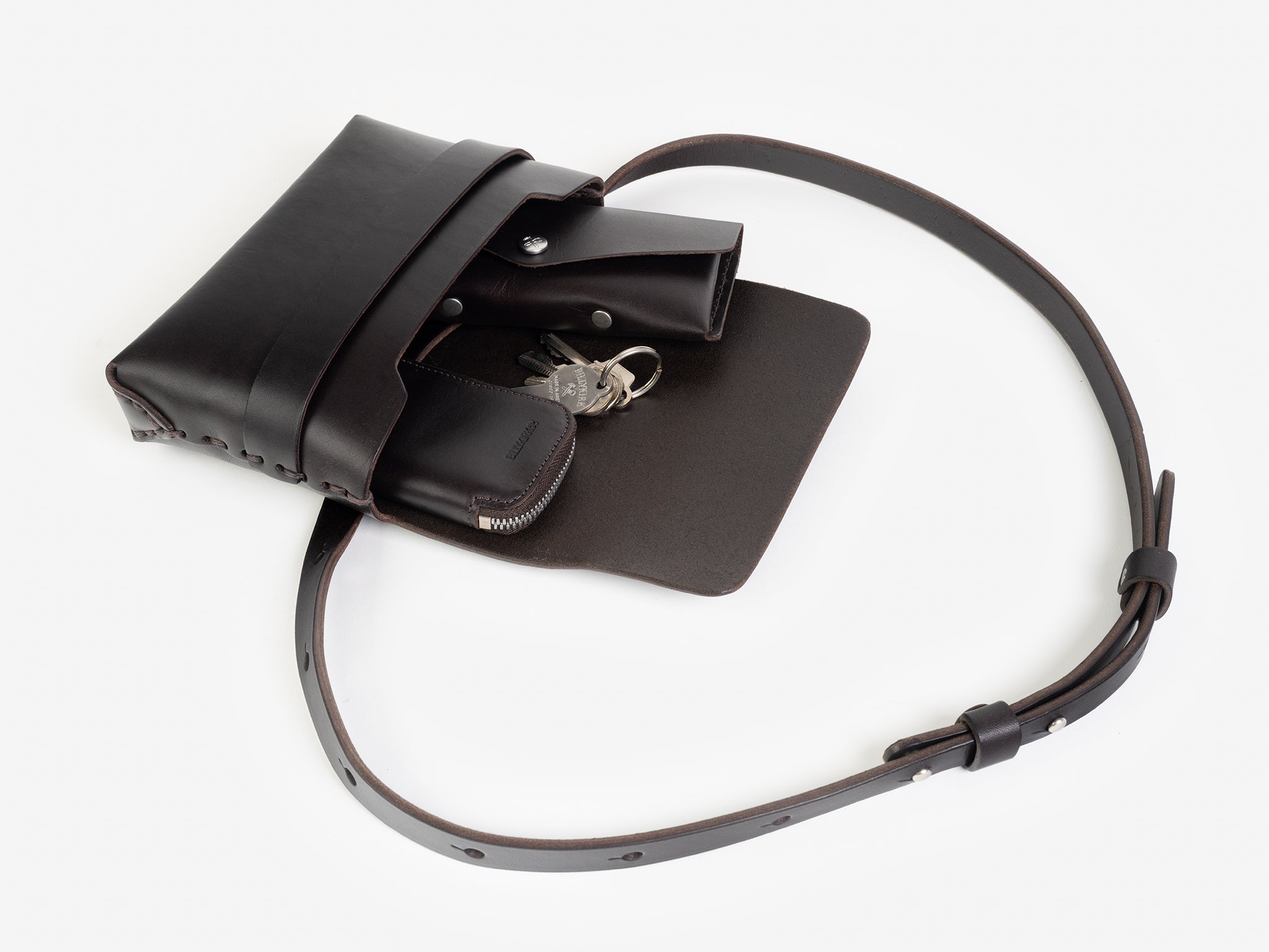 A dark brown leather crossbody bag with a flap open, revealing a zippered pouch a set of keys, and a leather sunglasses case inside. The bag has a long adjustable strap and minimalist design, displayed on a white background.