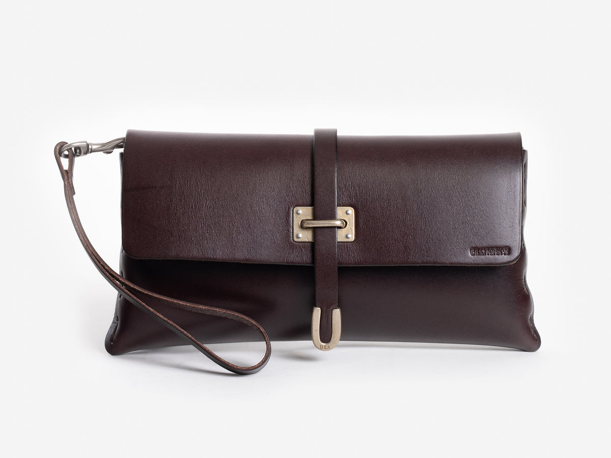 A dark brown leather clutch bag with a wrist strap, front flap closure, and a decorative brass buckle. The brand name is embossed on the lower right corner of the flap.