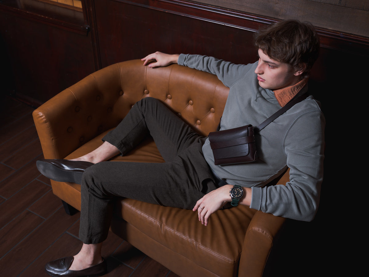 A young man lounges on a tan leather sofa, dressed in dark trousers, a gray sweatshirt, loafers, and a leather crossbody bag. He looks to the side with one arm resting on the sofa’s back. The room has dark wood flooring and walls.
