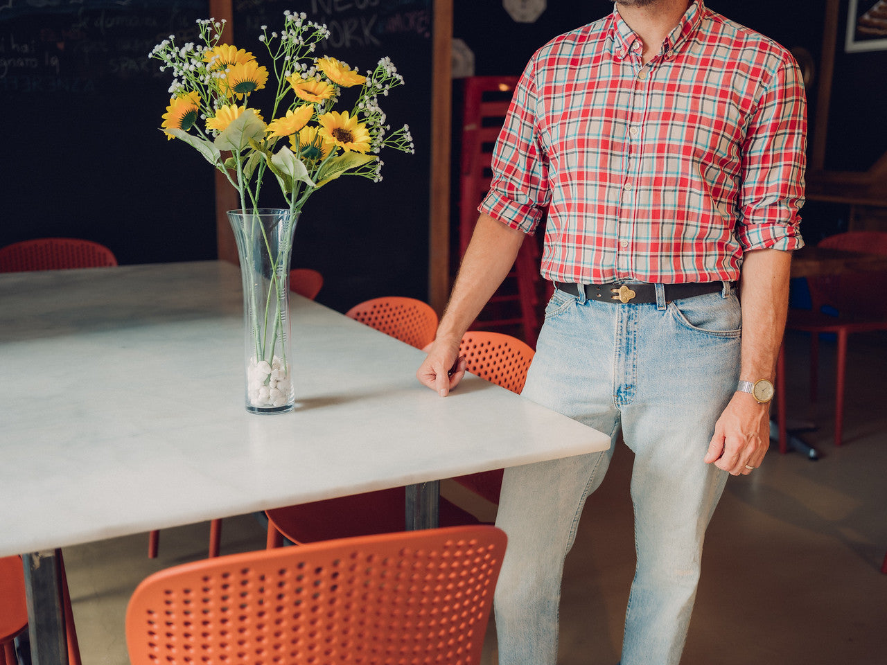 A person in a red plaid shirt and light jeans stands next to a white table with a tall vase of yellow flowers. Several orange chairs surround the table. The persons head is out of frame.