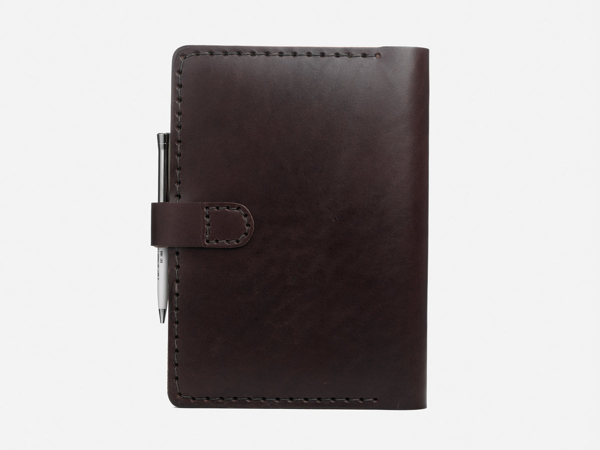 A dark brown leather notebook with visible stitching, a flap closure, and a silver pen attached to the left side, shown against a plain white background.