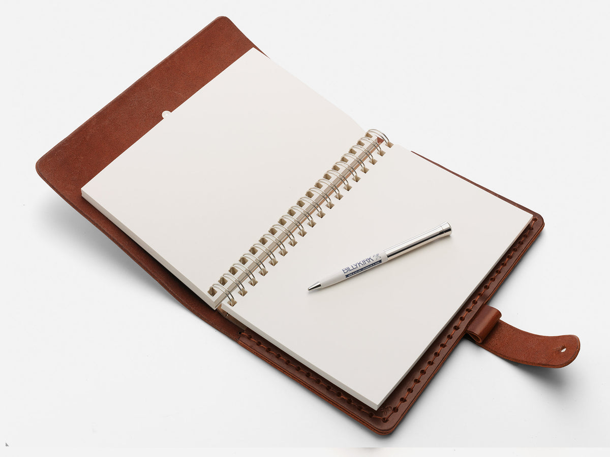 An open brown leather-bound notebook with blank white pages and a spiral binding lies on a white surface. A silver pen is resting diagonally across the pages.