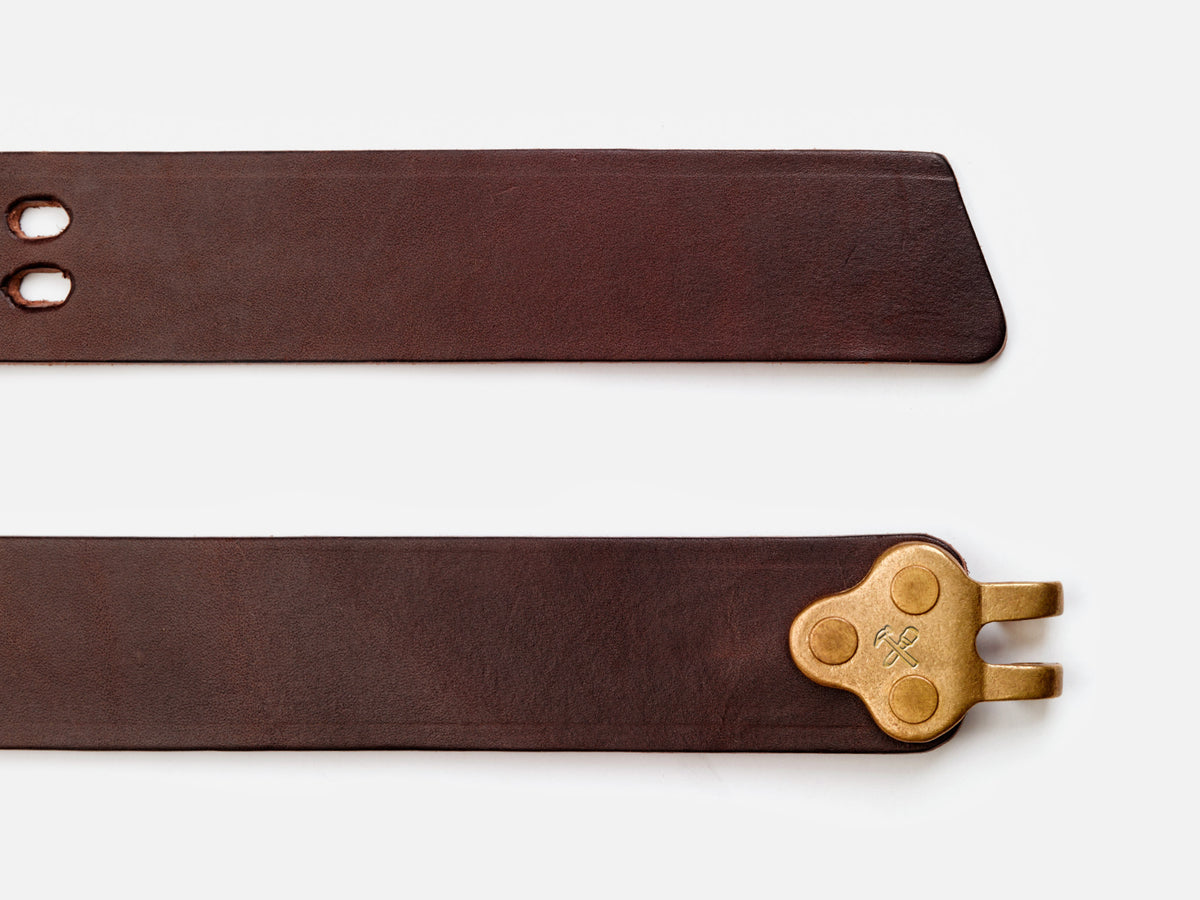 Two brown leather belt straps are shown. One end has two holes and a pointed tip, while the other end features a brass clasp with an engraved symbol. The background is white.