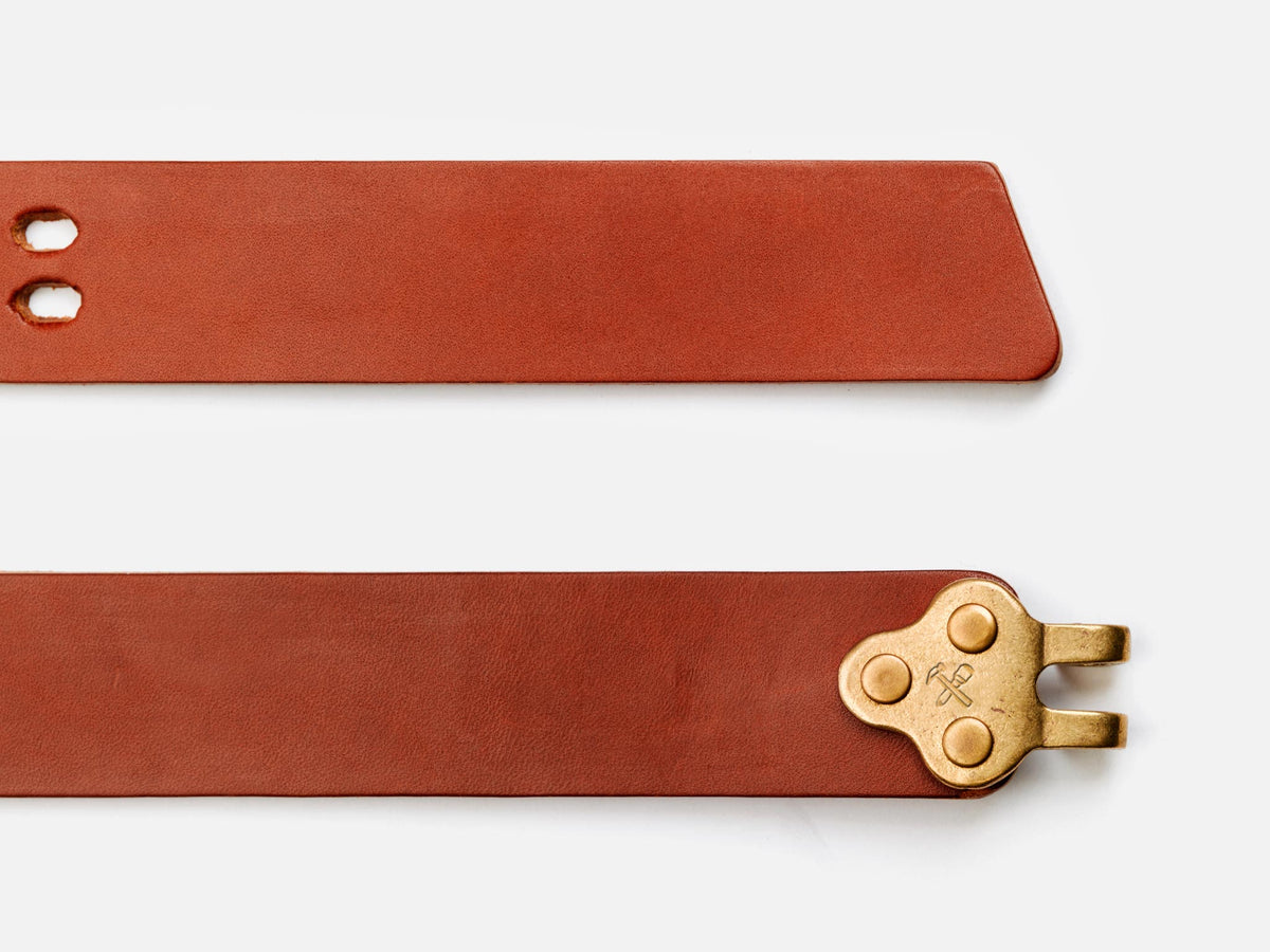 A brown leather belt shown flat, with one end featuring two holes and a pointed tip, and the other end displaying a brass buckle with three rivets.